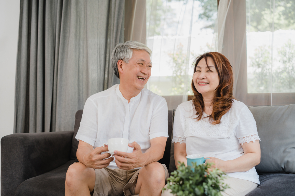 a senior man and a senior woman sit on a couch drinking coffee