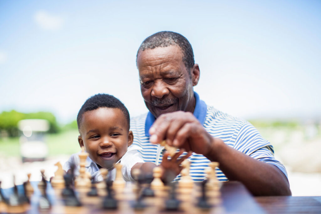 A senior man plays chess while holding his young grandson on his lap.