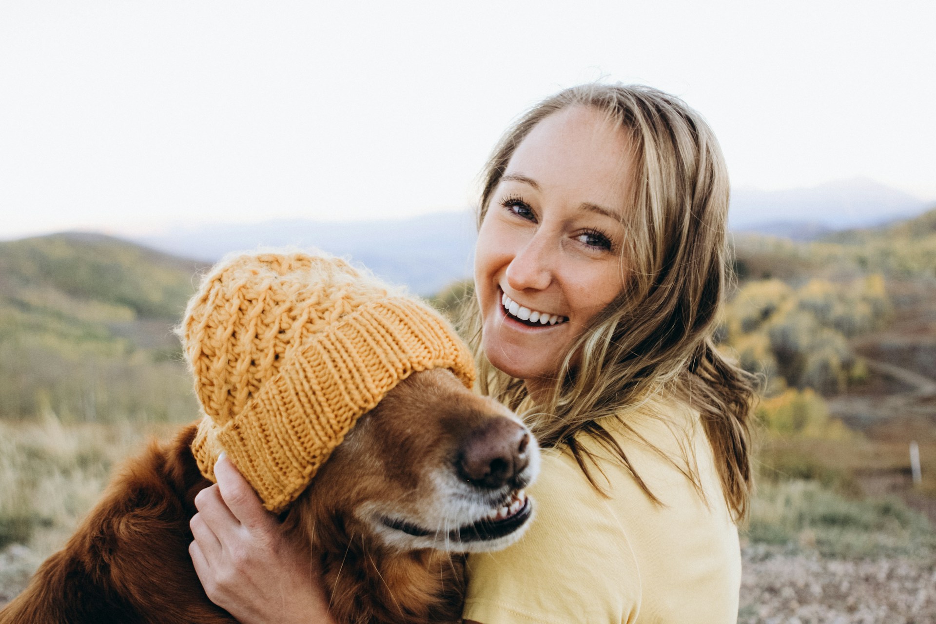 A woman puts a yellow knit hat on a dog and smiles at the camera
